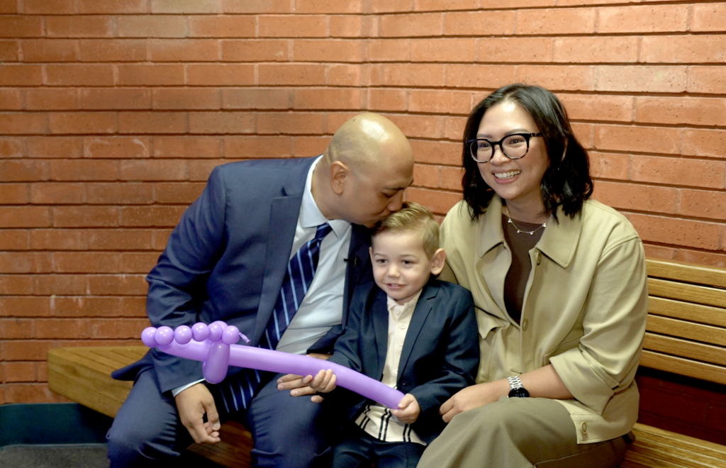 Family of three sitting on a bench. Father on the left, wearing a suit, leaning over kissing the top of his blond, adopted son's head. The son is holding a light purple balloon shaped as a sword. The mother, sitting and smiling on the right, is wearing a tan suit and eyeglasses.