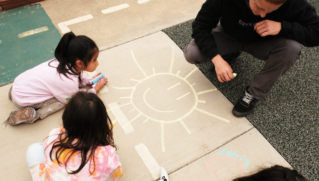 Children and an adult gathered around a chalk drawing of the sun on a sidewalk.